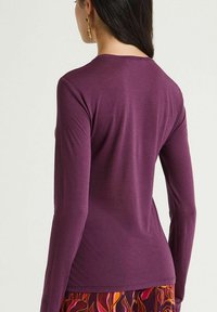 Maglietta a maniche lunghe aderente in tessuto color prugna scuro. Presenta una scollatura tonda e una texture liscia. Indossata con un capo inferiore a motivo.