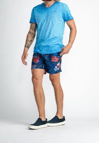Blå heatherad v-ringad t-shirt, blommiga marinblå badshorts med röda och rosa blommor, mörkblå slip-on skor, stående på en ljus bakgrund.