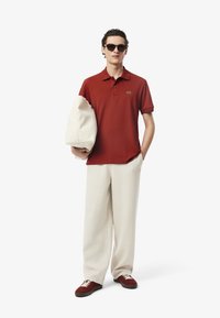 Röd polo med korta ärmar, beiga vida byxor och röda sneakers. Håller en vit väska. Innehåller Lacoste-logotypen på polon.