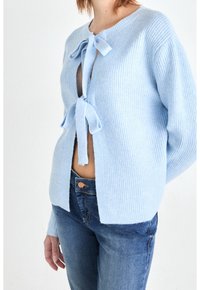 Cardigan tricoté bleu clair avec une fermeture à nouer, présentant une texture côtelée et un devant ouvert, associé à un jean bleu foncé.