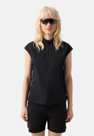 Blondynka ubrana w czarną bezrękawnikową bluzkę zapinaną z przodu na zamek, czarne szorty oraz duże ciemne okulary przeciwsłoneczne, stojąca na tle jednolitego jasnego tła.