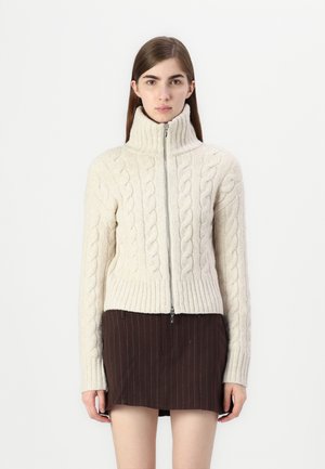 Cardigan crème en tricot torsadé avec un col montant et une fermeture éclair intégrale. Doté d'un ourlet et de poignets côtelés. Porté par-dessus une jupe sombre.