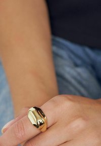 Guldtonad metallring med en slät, rundad design, med en reflekterande yta och en kupolform, bärs på en hand.