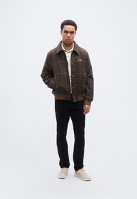 Καφέ σακάκι καρό με γιακά, κουμπιά κλικ και ριμπ μανσέτες, συνδυασμένο με λευκό πόλο πουκάμισο και μαύρο παντελόνι. Παπούτσια σε ανοιχτό μπεζ χρώμα.