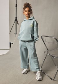 Vaaleansininen huppari edessä olevalla taskulla ja kirjotulla logolla, parina joustavat, väljät collegehousut. Kangas näyttää pehmeältä ja teksturoidulta.