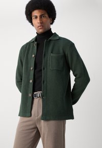 Chaqueta de camisa de color verde oscuro con textura, con cuello, cierre de botones y un bolsillo en el pecho, superpuesta sobre un suéter negro de cuello alto. Pantalones marrones.