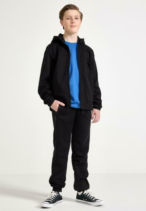 Teenagedreng står med hænderne i lommerne på sorte bukser, iført sort lynlåstrøje, blå t-shirt og sorte sneakers med hvide snørebånd.