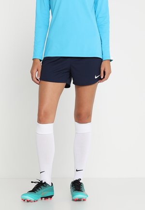 Dolna część ciała osoby noszącej jasnoniebieską koszulę z długim rękawem, granatowe spodenki z logo Nike, białe podkolanówki i turkusowe korki piłkarskie.