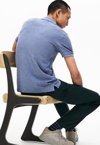 Kortärmad, ljusblå pikétröja med krage, kombinerad med mörkgröna byxor och ljusbeiga sneakers på en modern stol.