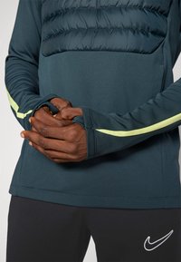 Mørk teal genser med quiltet overdel, tettsittende mansjetter med lysegule striper og en glatt tekstur. Nike-logo på buksene.