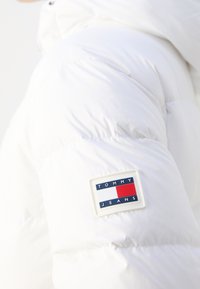 Gros plan sur la manche d'une doudoune blanche avec un patch logo rectangulaire Tommy Jeans en bleu marine, blanc et rouge.