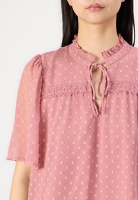Blouse rose avec une texture à pois, col volanté, décolleté en trou de serrure et manches courtes bouffantes, avec des détails en dentelle sur la poitrine.