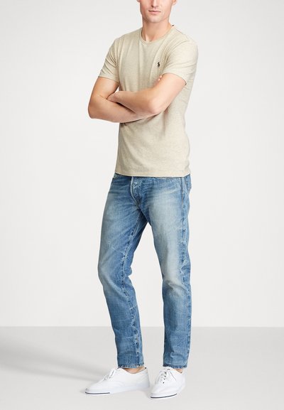 Beige cotton T-shirt with round neckline, paired with light blue denim jeans. White sneakers complete the outfit. Man stands with arms crossed.