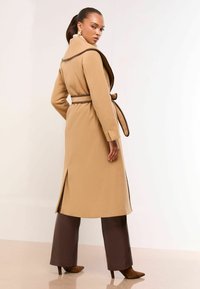 Beige langer Mantel mit einem Taillengürtel, featuring einen Schalkragen und braunen Akzenten. Das Design hat einen Rücken-Schlitz und eine glatte Textur, kombiniert mit braunen Hosen.