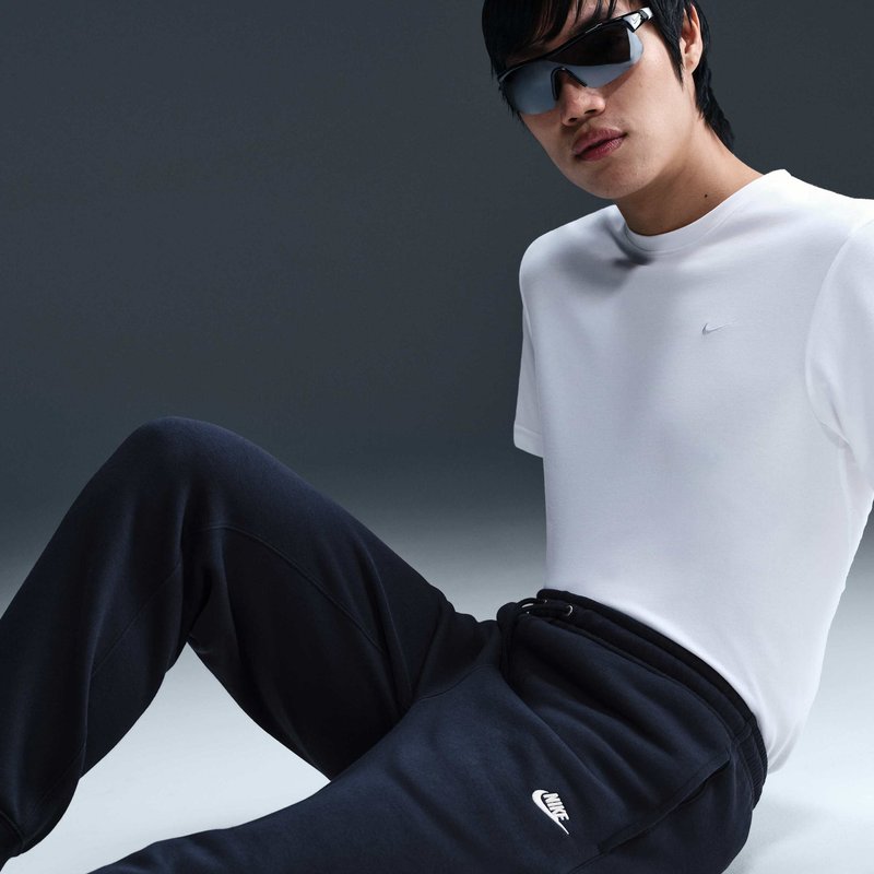Persona che indossa occhiali da sole neri, una maglietta Nike bianca e pantaloni della tuta Nike neri, seduta in posa rilassata su uno sfondo semplice.