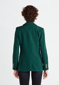 Personne aux cheveux courts et bouclés portant un blazer vert foncé cintré et un pantalon noir, vue de dos sur un fond blanc.