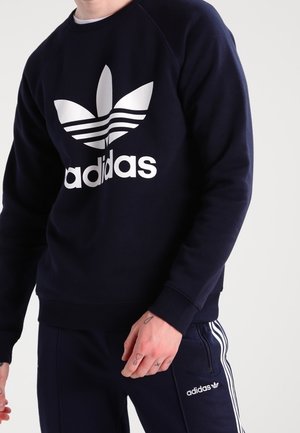 Personne portant un sweat Adidas bleu marine avec grand logo trèfle blanc et un pantalon de survêtement Adidas assorti avec des bandes blanches et logo sur la cuisse.