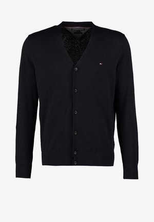 Hombre caminando hacia adelante llevando un cárdigan negro, camisa blanca, vaqueros oscuros, zapatos negros y un bolso marrón cruzado.