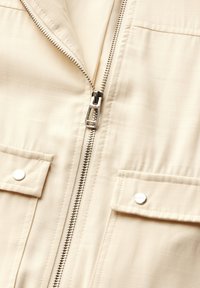 Beige zip-up jacka i ett slätt, lättviktigt material. Har silverdetaljer, två fickor med tryckknappar och en minimalistisk design.