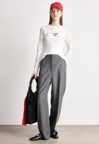 Cămașă albă cu mânecă lungă, cu text, pantaloni largi gri cu dungi, geantă de umăr neagră, cască roșie și pantofi negri cu tinte.