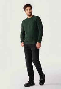 Pull en maille côtelée vert foncé avec un col rond, associé à un jean noir et des bottes noires à lacets, avec des poignets et un ourlet côtelés.