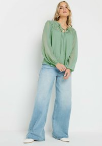 Blusa leggera verde chiaro, trasparente, con pois, colletto con lacci e dettagli arricciati, abbinata a jeans blu a vita alta e gamba larga e scarpe bianche.