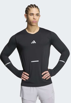 Man met gevlochten haar die een zwart lange-mouwen sportshirt met reflecterende details en lichtgrijze shorts draagt, handen in de heupen.
