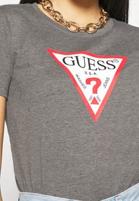 Šedé bavlněné tričko s velkým červeným trojúhelníkovým grafickým vzorem, na kterém je slovo "GUESS" spolu s otazníkem a brandingovými detaily.