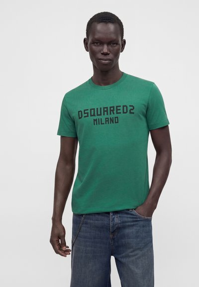 Άνδρας που φοράει πράσινο μπλουζάκι Dsquared2 Milano και μπλε τζιν με το ένα χέρι στην τσέπη, μπροστά από απλό φόντο.