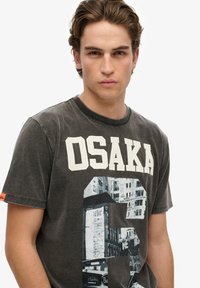 La camiseta de algodón gris presenta una gran impresión de 'OSAKA' en blanco y un gráfico negro de una escena urbana. Cuello redondo clásico con mangas cortas.
