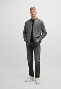 Camisa de botón gris con un bolsillo, usada sobre una camiseta negra, combinada con jeans oscuros y zapatillas blancas. Tela suave con ligera textura.