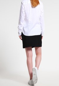 Camisa blanca de manga larga con pequeños lunares coloridos, combinada con una falda negra y zapatillas blancas, vista desde la parte de atrás.