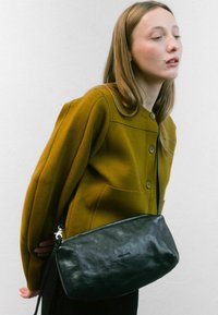 Borsa in pelle nera con una texture liscia e chiusura a zip, dotata di una tracolla lunga. Indossata da un modello con un cardigan color senape.