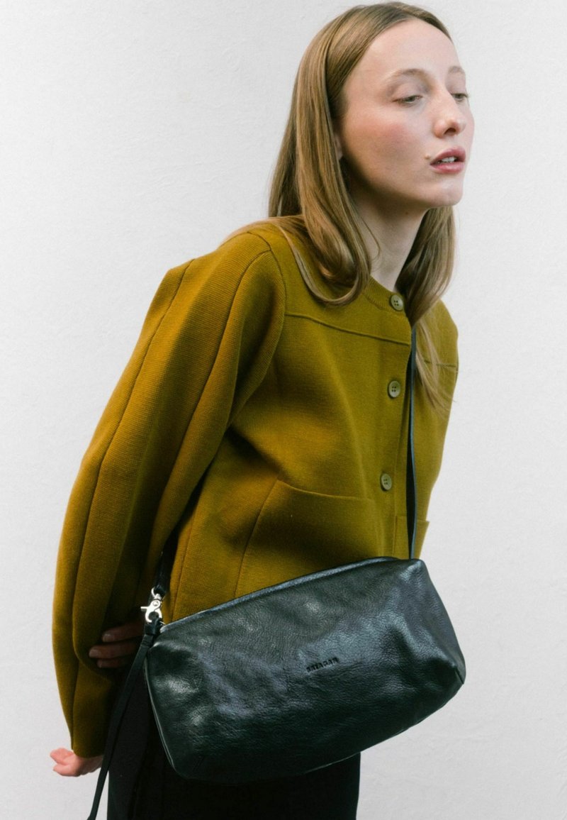 Borsa in pelle nera con una texture liscia e chiusura a zip, dotata di una tracolla lunga. Indossata da un modello con un cardigan color senape.