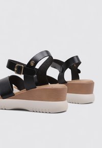 Sandalias de cuero negro con dos correas ajustables, un hebilla dorada y una suela en cuña en beige y crema. Suela de goma texturizada para mayor agarre.