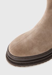 Beige sælskindsstøvle med syet søm, sort lagdelt sål og struktureret greb på en hvid baggrund.
