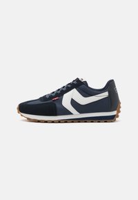 Chaussures de sport bleu marine avec tige en daim et nylon, motif vague blanc, semelle extérieure en gomme texturée et lacets gris. Détail de logo rouge sur le côté.