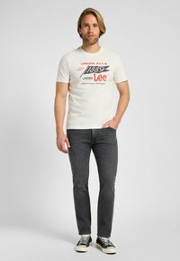Camiseta blanca de manga corta con el logo "Lee" y un diseño de texto vintage, combinada con jeans slim-fit en gris oscuro y zapatillas negras con suelas blancas.