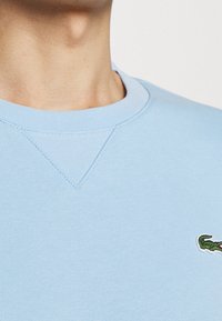 Ljusblå bomulls sweatshirt med klassisk rund hals, med en liten grön krokodil-logotyp och strukturerade sömmar.