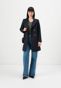 Femme debout dans un manteau croisé bleu marine, un haut vert en V, un jean large bleu, des chaussures à talons noires et portant un petit sac à épaule noir.