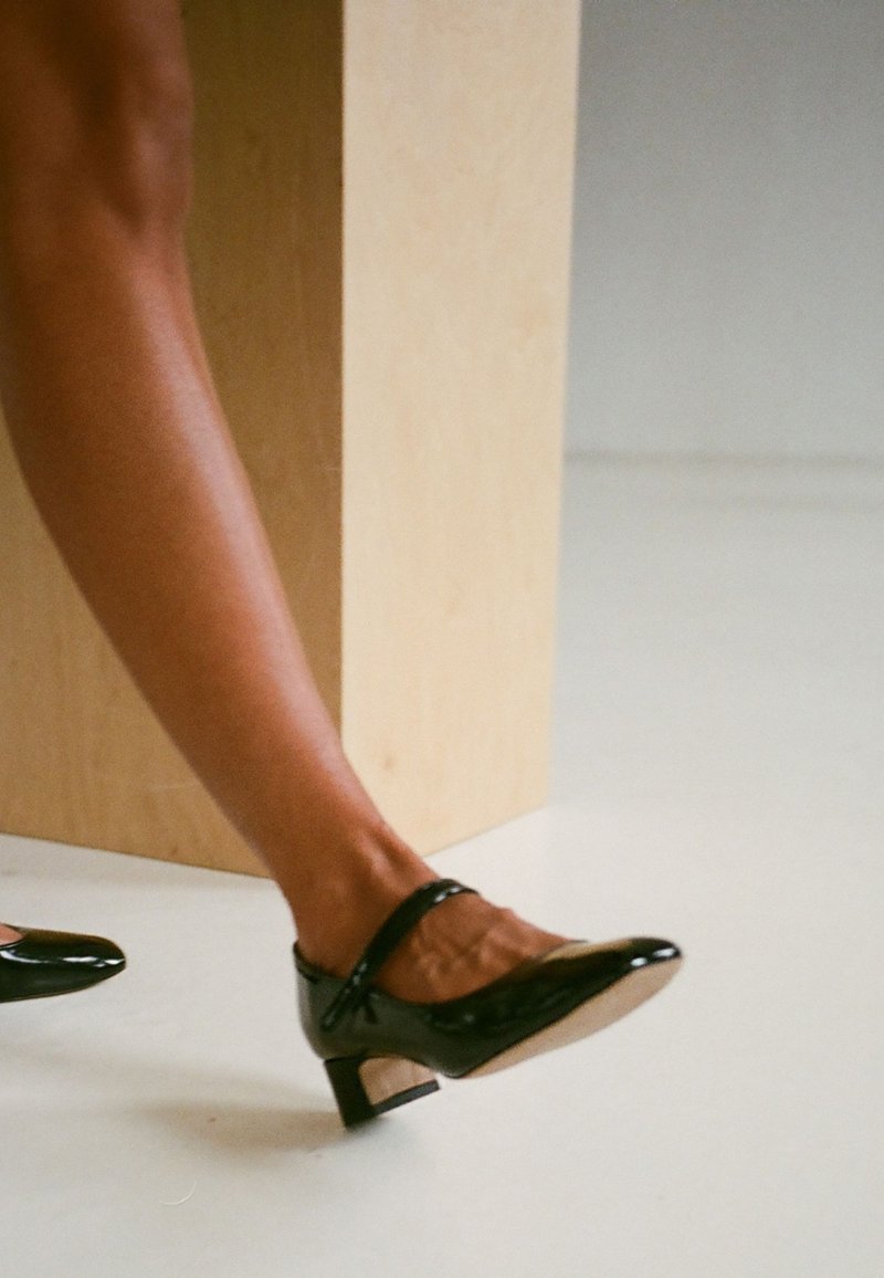 Scarpe low-heel in pelle verniciata nera con punta affusolata e cinturino alla caviglia; tacco a blocco in legno; superficie liscia con tagli superficiali.