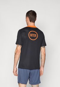Svart idrottströja med orange krage och "INTER"-logotyp. Korta ärmar, lättviktsmaterial och en bekväm passform. Bärs tillsammans med marinblå shorts.