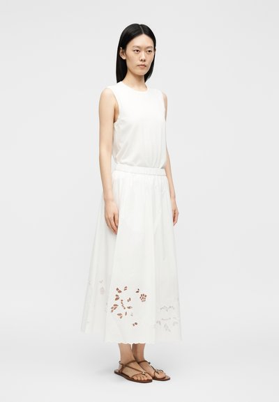 Kvinde med langt sort hår iført en hvid ærmeløs top, hvid midi nederdel med udskårne blomster-dekorationer og brune flade sandaler står med fronten mod kameraet.