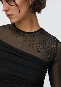 Musta mekko, jossa on läpinäkyvä verkkopäällinen koristeltu mustilla helmillä ja kerätty kangassuunnittelu, joka luo tekstuurisen ilmeen.