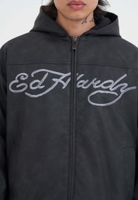 Giovane che indossa una felpa nera con zip e con una grande scritta grigia "Ed Hardy" sul petto, con un tatuaggio sul collo parzialmente visibile.