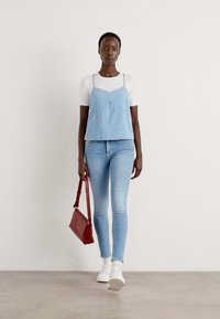 Denim camisole over a white T-shirt, paired with light blue skinny jeans and white sneakers. A small red handbag completes the outfit.