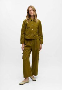Giacca cropped color verde oliva con due tasche frontali e chiusura con bottoni, abbinata a pantaloni cargo a gamba larga dello stesso colore e scarpe flat beige chiaro.
