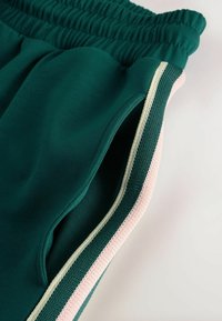 Grüne Sportshorts mit einem elastischen Bund und zwei Seitentaschen. Akzente bestehen aus cremefarbenen und pinken Streifen entlang der Kanten.