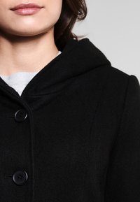 Abrigo de lana negro con capucha, con cierres de botones y una textura suave. Detalle de costura visible en el cuello y la costura del hombro.