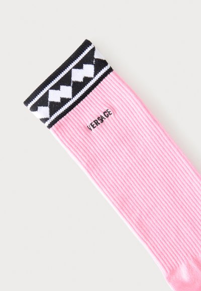 Chaussette côtelée rose avec motif géométrique noir et blanc au niveau du bord-côte et logo "VERSACE" brodé en noir près du haut.